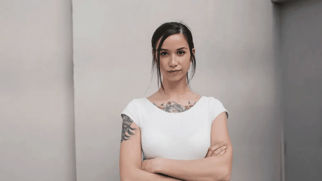 Woman with tattoos wearing a white top standing against a plain background